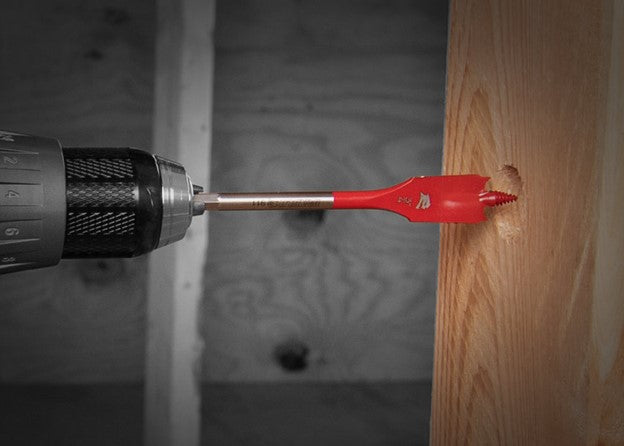 A red spade drill bit from the Diablo DIABLO DSP1910-S3 3-Piece Spade Bit Set for Wood is shown attached to a power drill, efficiently drilling a hole into a wooden board. The background features a blurred gray surface, highlighting the precision and reliability of Diablo's craftsmanship in action.