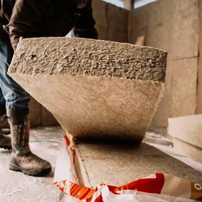 A person in boots is handling a large ROCKWOOL RXCB381525 R38 ComfortBatt insulation panel, made of rock-based mineral fibers, inside a building under construction. The sustainable insulation panel is lifted from a partially opened package on the floor.