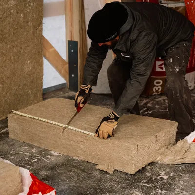 In a construction area, a worker in gear measures and cuts sustainable ROCKWOOL RXCB381525 R38 ComfortBatt Insulation, sized at 9.5x15-1/4x47 (19.9 SQF).