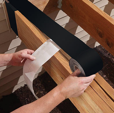 Using Protecto Wrap Deck Joist Flashing Tape, a person unrolls and positions it along a wooden beam, utilizing Super Stick Technology for better moisture protection, set against a backdrop of siding walls.