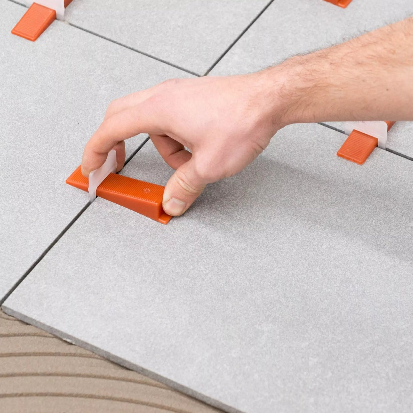 A hand expertly uses RAIMONDI 180CUNEO500 RLS Level Wedges in orange on gray tiles, showcasing a seamless setting process. Available as a 500-pack at Leroy Merlin for $64.99.