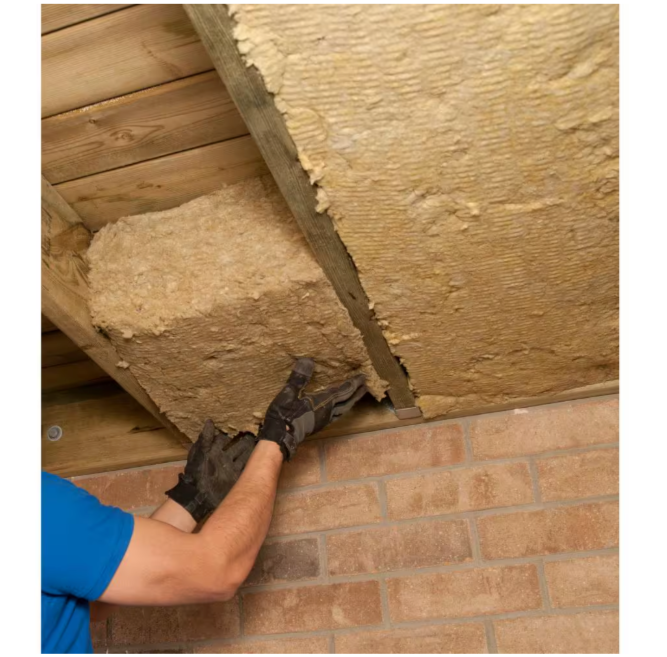 A person in a blue shirt and gloves installs Rockwools ROCKWOOL ComfortBatt R-23 Stone Wool Insulation (5-1/2 in. x 15.25 in. x 47 in.) under a wooden ceiling, fitting large beige pieces between beams to enhance thermal performance over the brick wall.