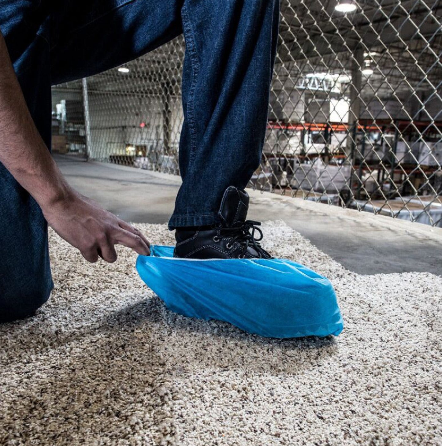 In a well-lit warehouse by the chain-link fence, someone kneels on gravel and slips a SURFACE SHIELDS SC3001PB blue disposable shoe cover over their black boot. These waterproof covers from SURFACE SHIELDS offer protection and slip resistance.