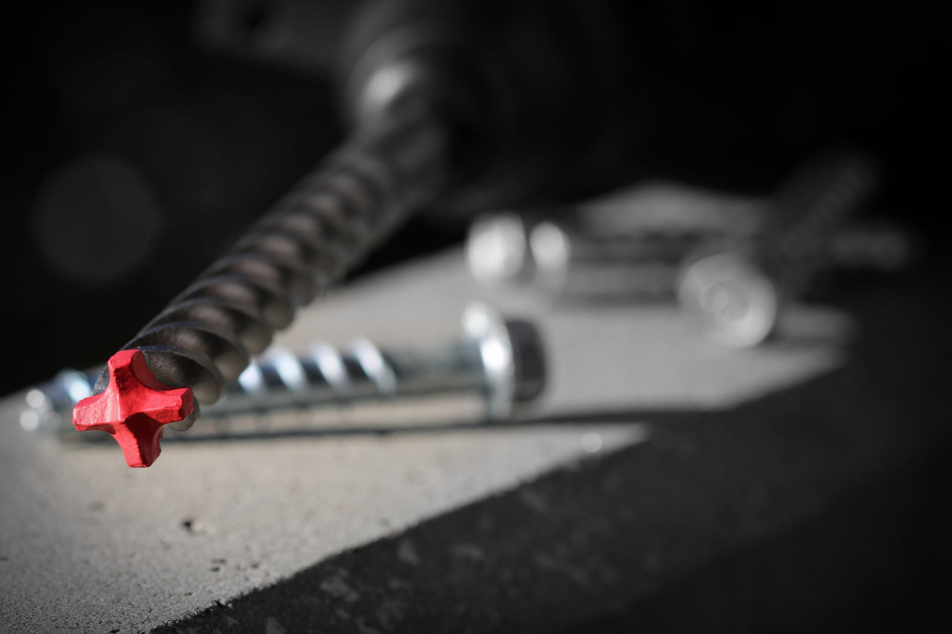 Close-up of a DIABLO DMAPL4332 Rebar Demon drill bit with a red tip on concrete, surrounded by metal screws. The blurred background highlights the focused Diablo 7/16 in. x 6 in. x 8 in. SDS-Plus Hammer Drill Bits presence in the foreground.