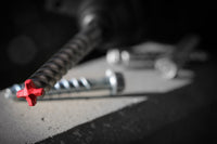 Close-up of a DIABLO DMAPL4332 Rebar Demon drill bit with a red tip on concrete, surrounded by metal screws. The blurred background highlights the focused Diablo 7/16 in. x 6 in. x 8 in. SDS-Plus Hammer Drill Bits presence in the foreground.