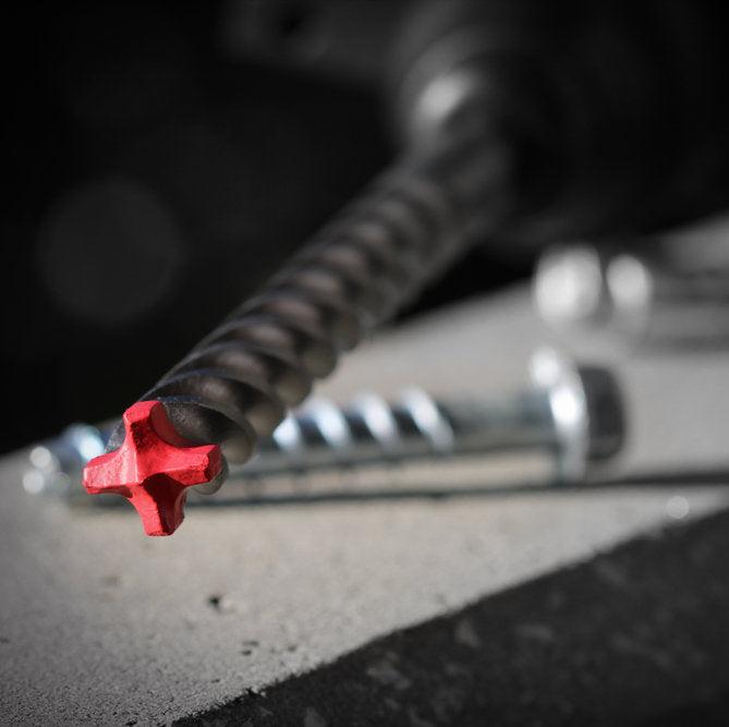 A close-up of a Diablo Rebar Demon™ SDS-Plus drill bit with a red tip on a gray surface, background shows two slightly blurred parallel screws.