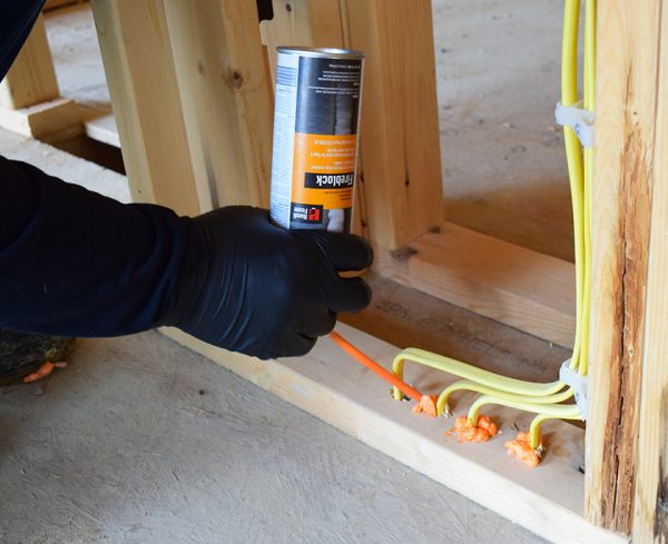 A person in black gloves applies HANDIFOAMs orange P30033 FireBlock Spray Foam Sealant to gaps around electrical wiring in a wooden frame. The silver can features text, and the scene occurs at a construction site focused on preventing flame spread.