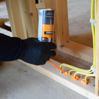 A person in black gloves applies HANDIFOAMs orange P30033 FireBlock Spray Foam Sealant to gaps around electrical wiring in a wooden frame. The silver can features text, and the scene occurs at a construction site focused on preventing flame spread.