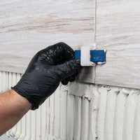 A person wearing a black glove is using a blue tool to adjust the spacing between two beige tiles, secured with MAPEI Keraflex SG Large-and-Heavy-Tile Mortar with Polymer by Mapei. The scene suggests a tiling installation in progress, with the white adhesive clearly visible beneath the tiles.