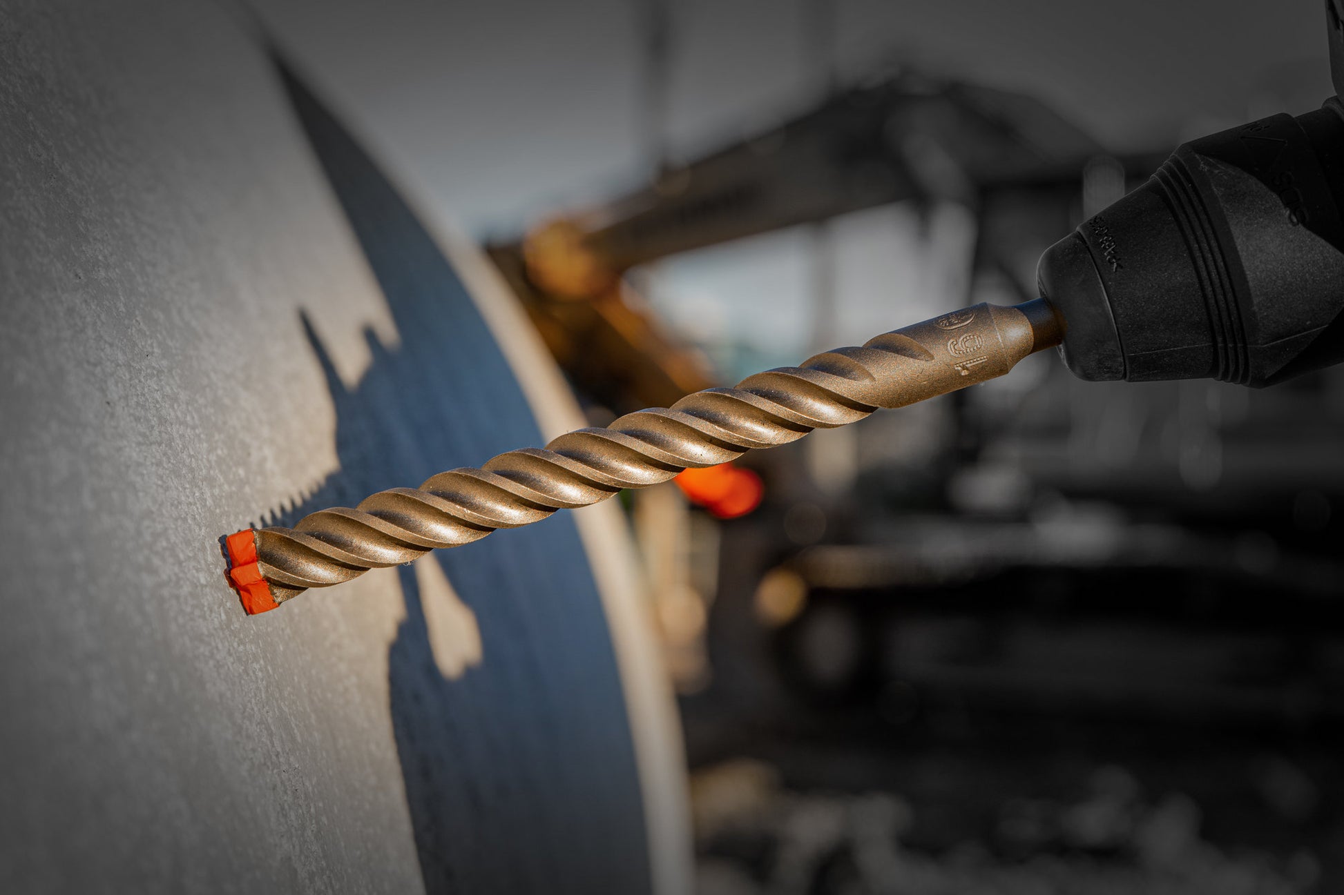 Close-up of a DIABLO DMAPL4332 Rebar Demon™ SDS-Plus bit drilling into concrete. Sunlight casts a shadow on the wall, while blurred construction machinery emphasizes the precision Diablo guarantees.