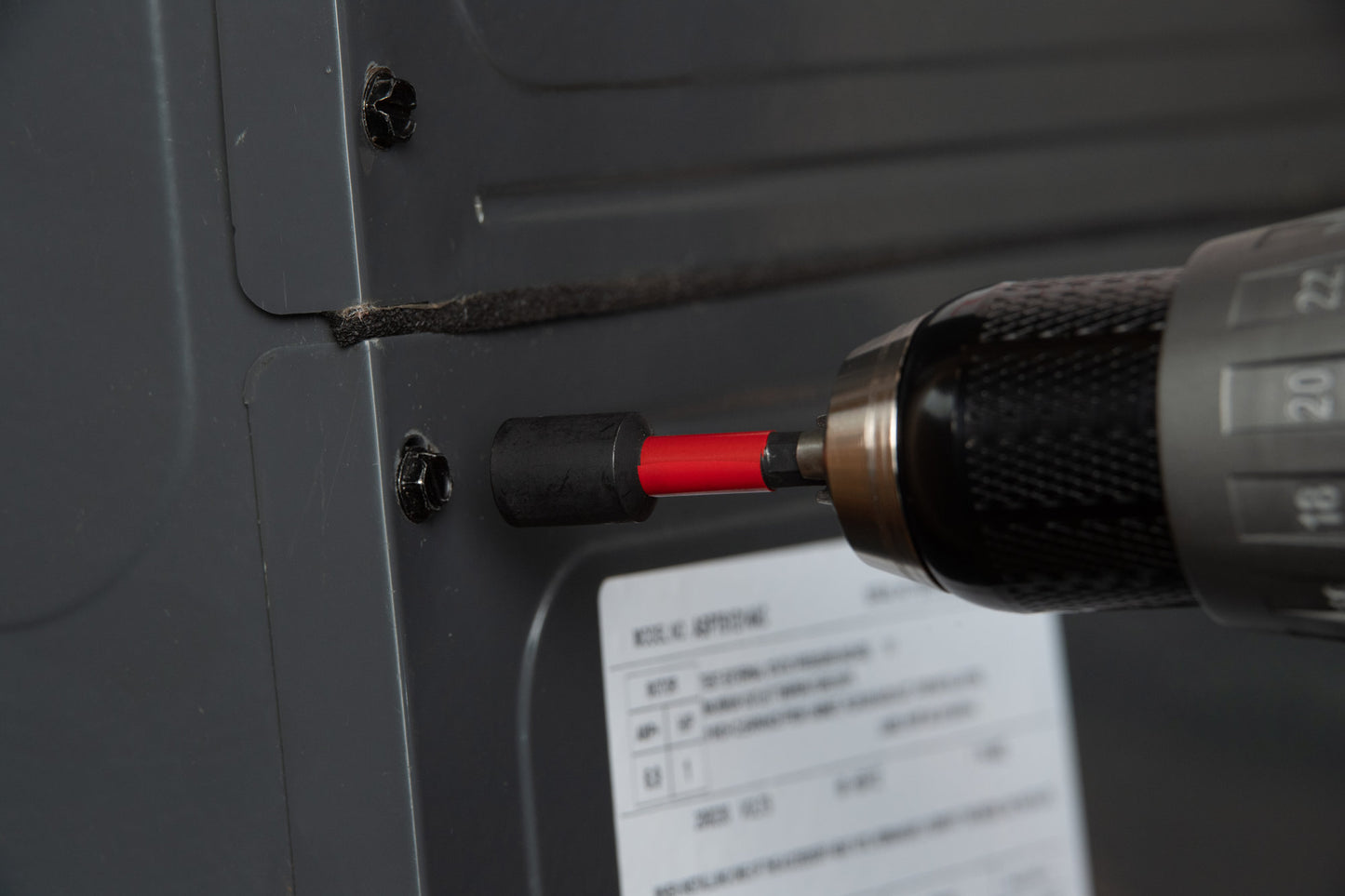 A close-up of a Diablo DIABLO DNTSL-S5 2-9/16 in. Magnetic Nut Driver with a red and black bit using Impact Strong™ technology tightens a screw on a gray metal panel. The panel has a white label, text, and symbols, with dust visible around the screws.