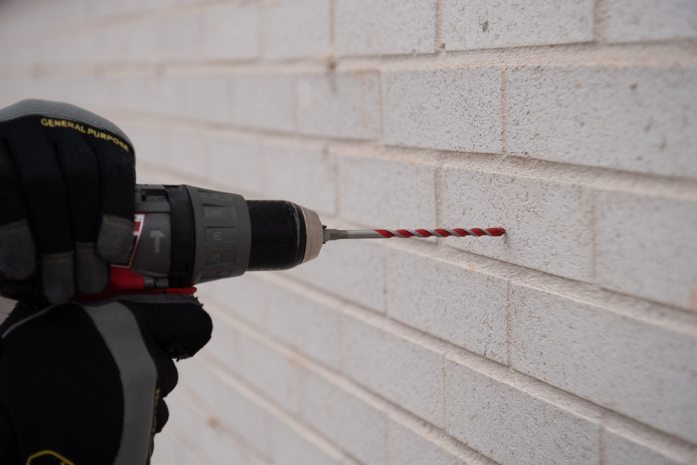 A gloved person uses an Impact Strong™ power drill with a Diablo SPEEDemon™ red granite carbide-tipped hammer drill bit for precision drilling into a white brick wall.