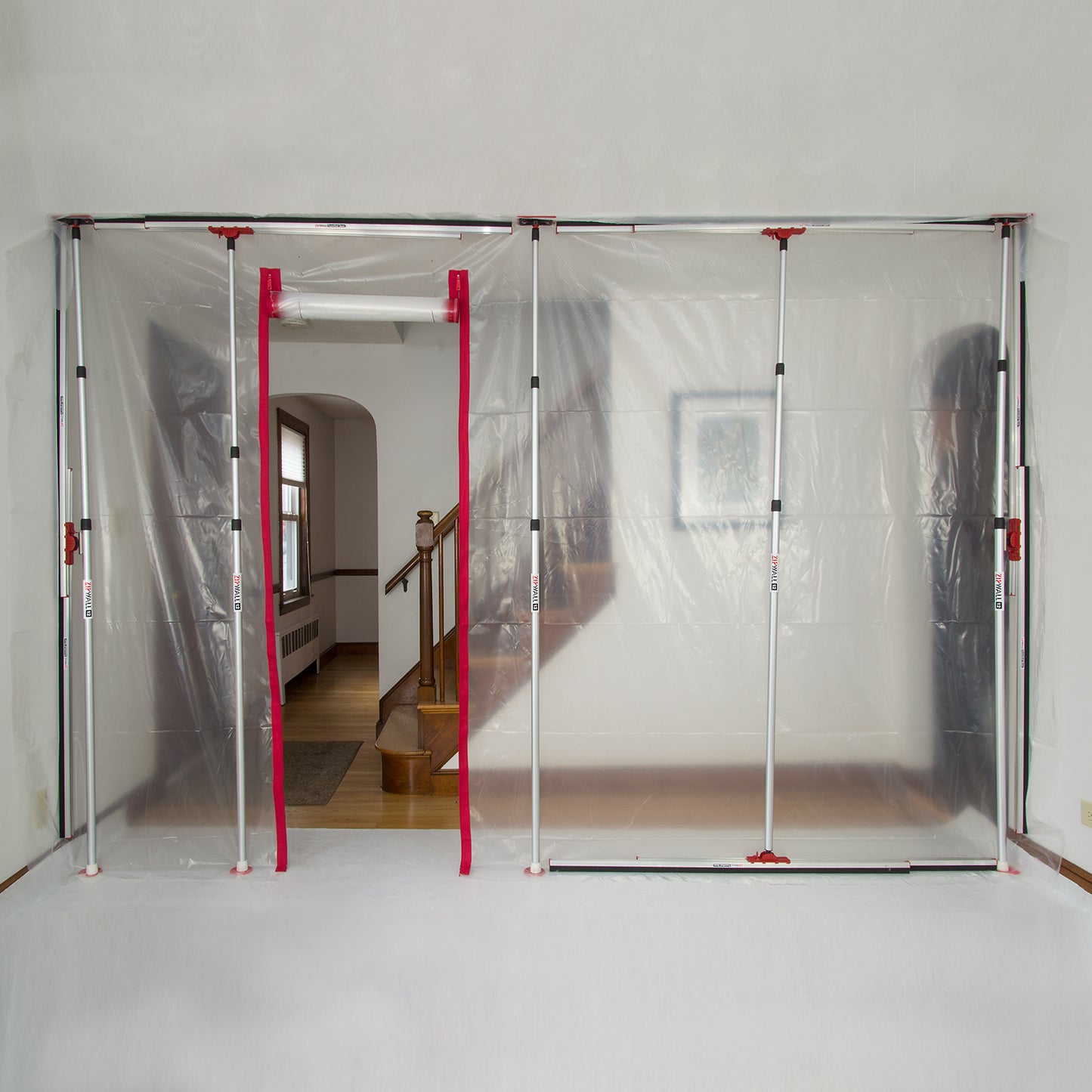 A ZIPWALL HDAZ2 barrier with heavy-duty zippers elegantly divides a room with wooden stairs and a framed picture, supported securely by four adjustable poles.